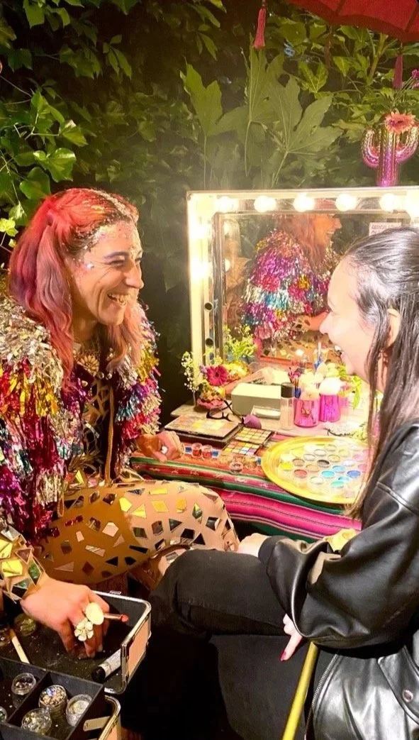 Two women at a makeup vanity, smiling and laughing, with colorful clothing and glitter on one woman's face, surrounded by makeup and decorative items in an outdoor setting with greenery.