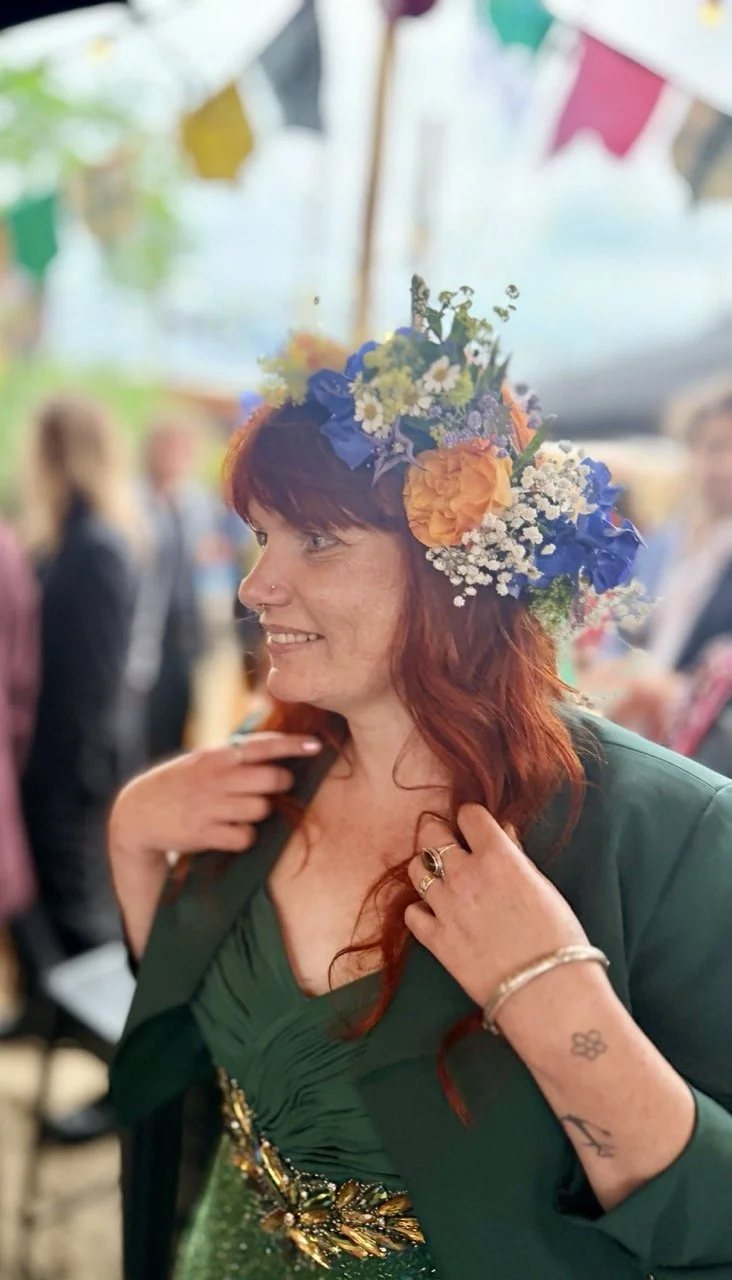 Woman with red hair wearing a green dress and a flower crown at a festive outdoor event.