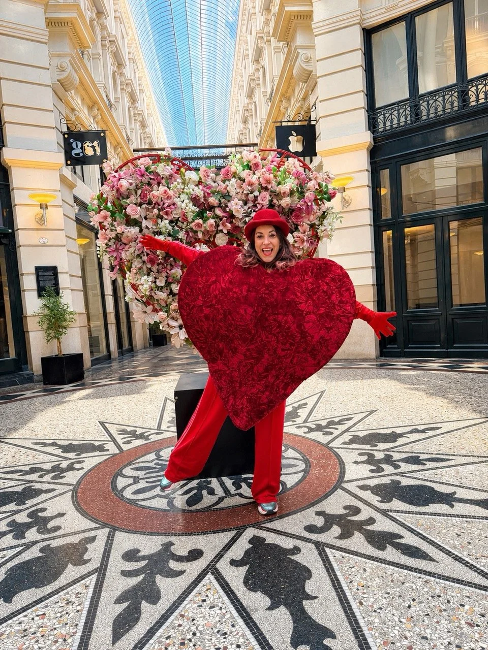 Woman dressed in red with a heart-shaped floral costume, standing in a shopping mall with a large flower arrangement and decorative flooring.