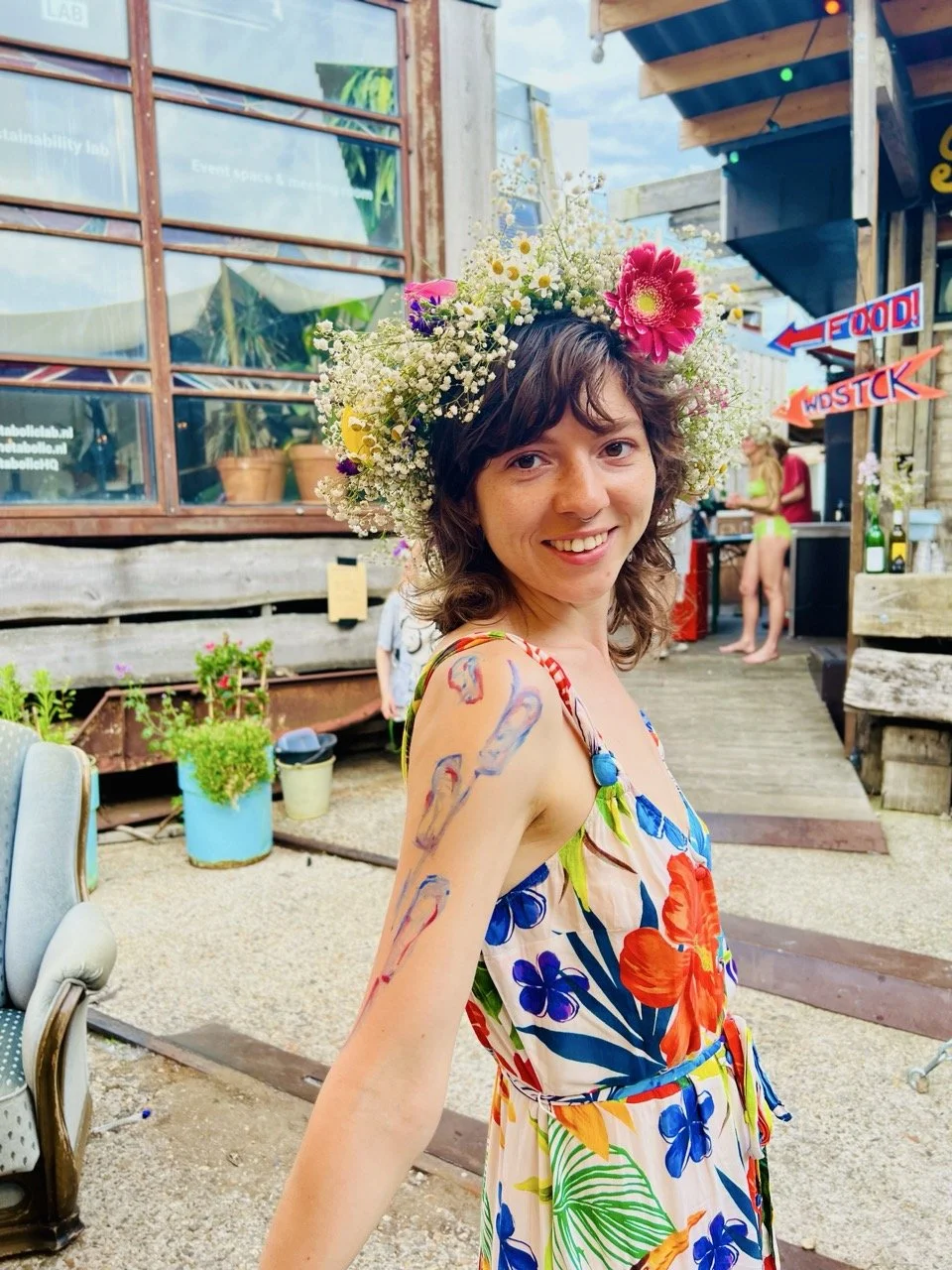 A woman with shoulder-length brown hair wears a floral dress and a large flower crown, smiling outdoors amidst colorful signs and plants.
