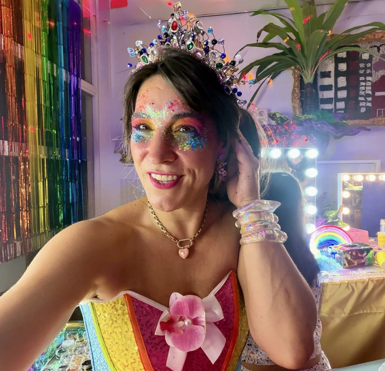 A woman wearing a birthday crown and rainbow-colored dress with a pink flower on the front, smiling at a lively party with rainbow decorations, colorful makeup with glitter and sequins, and a rainbow-themed cake in the background.