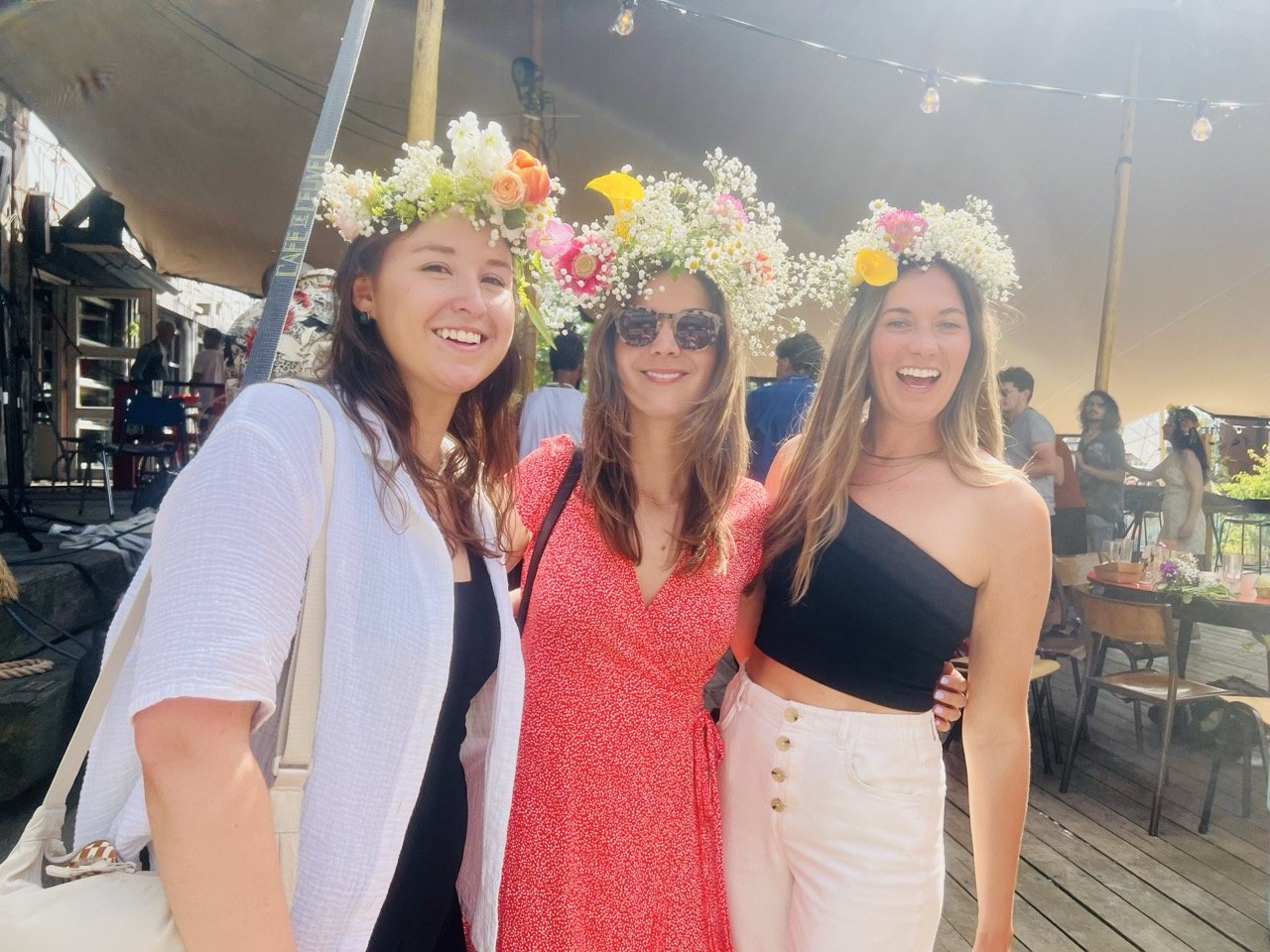 Three women wearing floral crowns made of baby's breath and colorful flowers smiling at the camera at an outdoor event.