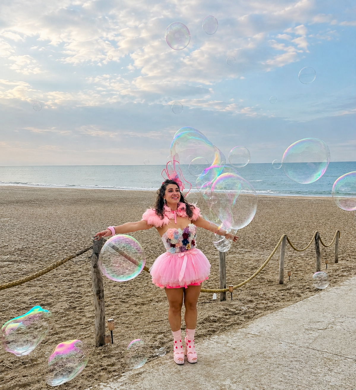 Woman dressed in a pink fairy costume with a tutu and wings, standing on a sandy beach, surrounded by floating soap bubbles, with the ocean and cloudy sky in the background.