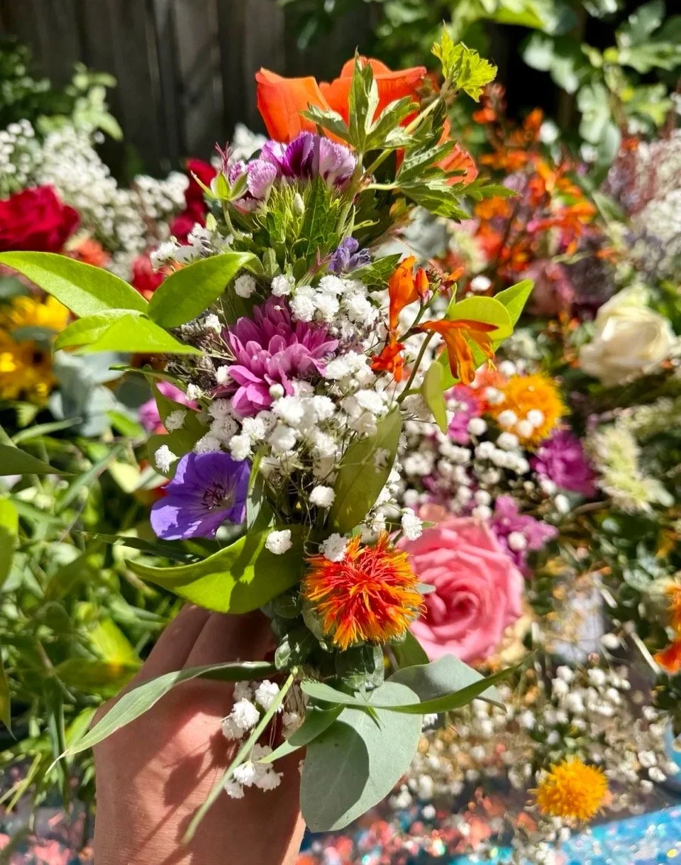 A colorful bouquet of various flowers including pink roses, orange and yellow blossoms, purple and white flowers, and greenery, held in a person's hand with a blurred garden background.