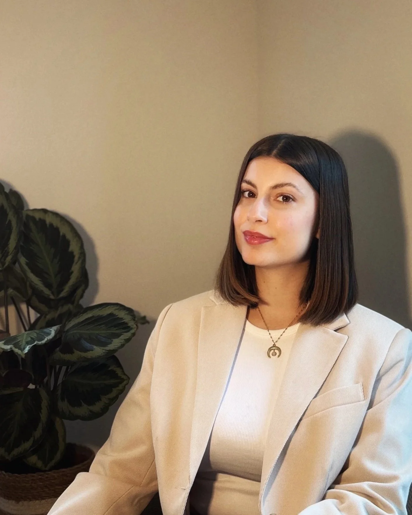 A woman with shoulder-length dark hair, wearing a white top and beige blazer, sitting next to a large leafy plant, with a neutral colored wall in the background.
