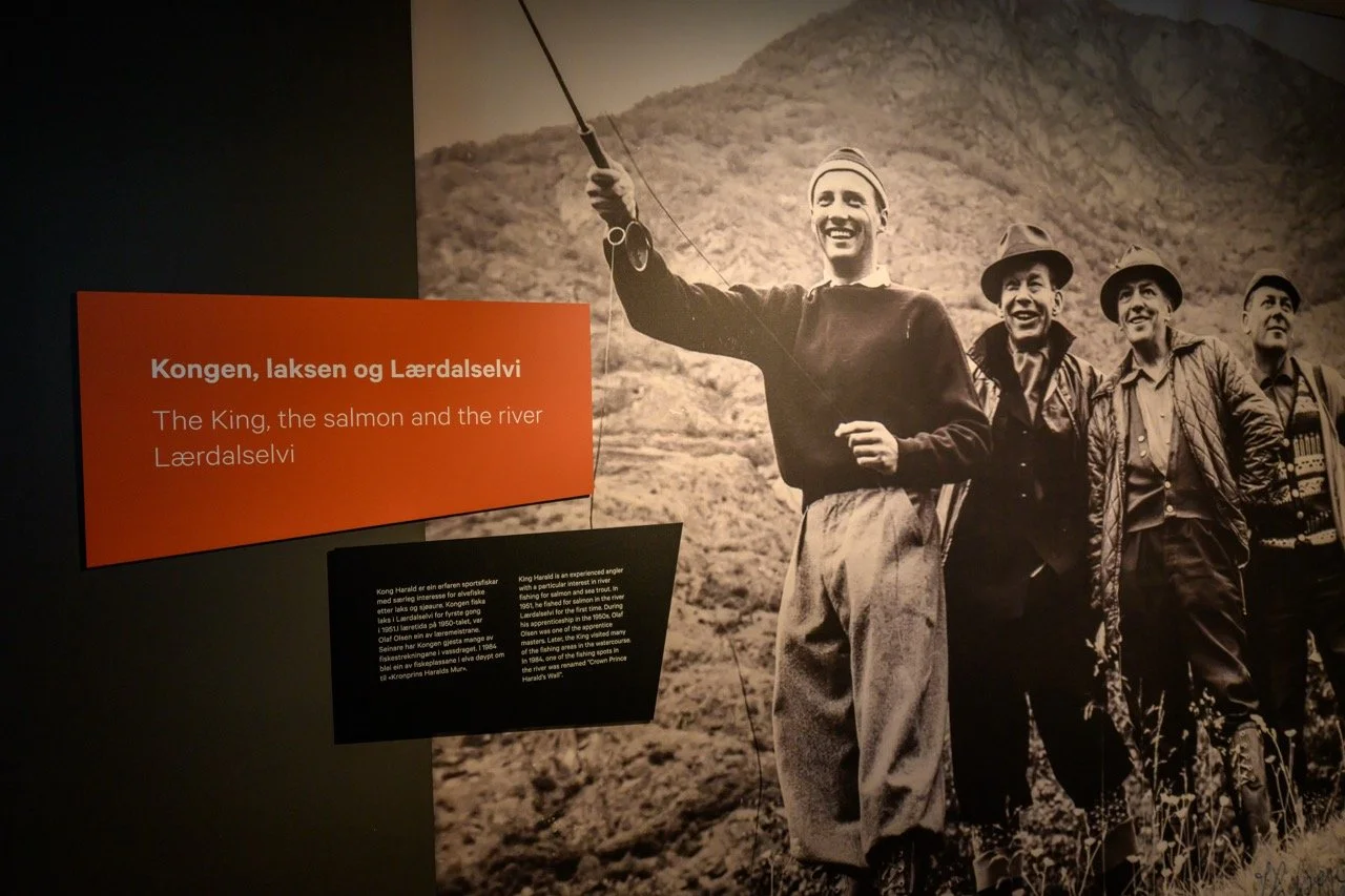 Historisk svart-hvitt fotografi av fire personer som går langs en fjellside, hvorav en holder en fiskestang. En av personene holder opp en fisk. Det er en informasjonstavle med tekst på norsk og engelsk ved siden av fotoet. Temem: Kongen, laksen og L