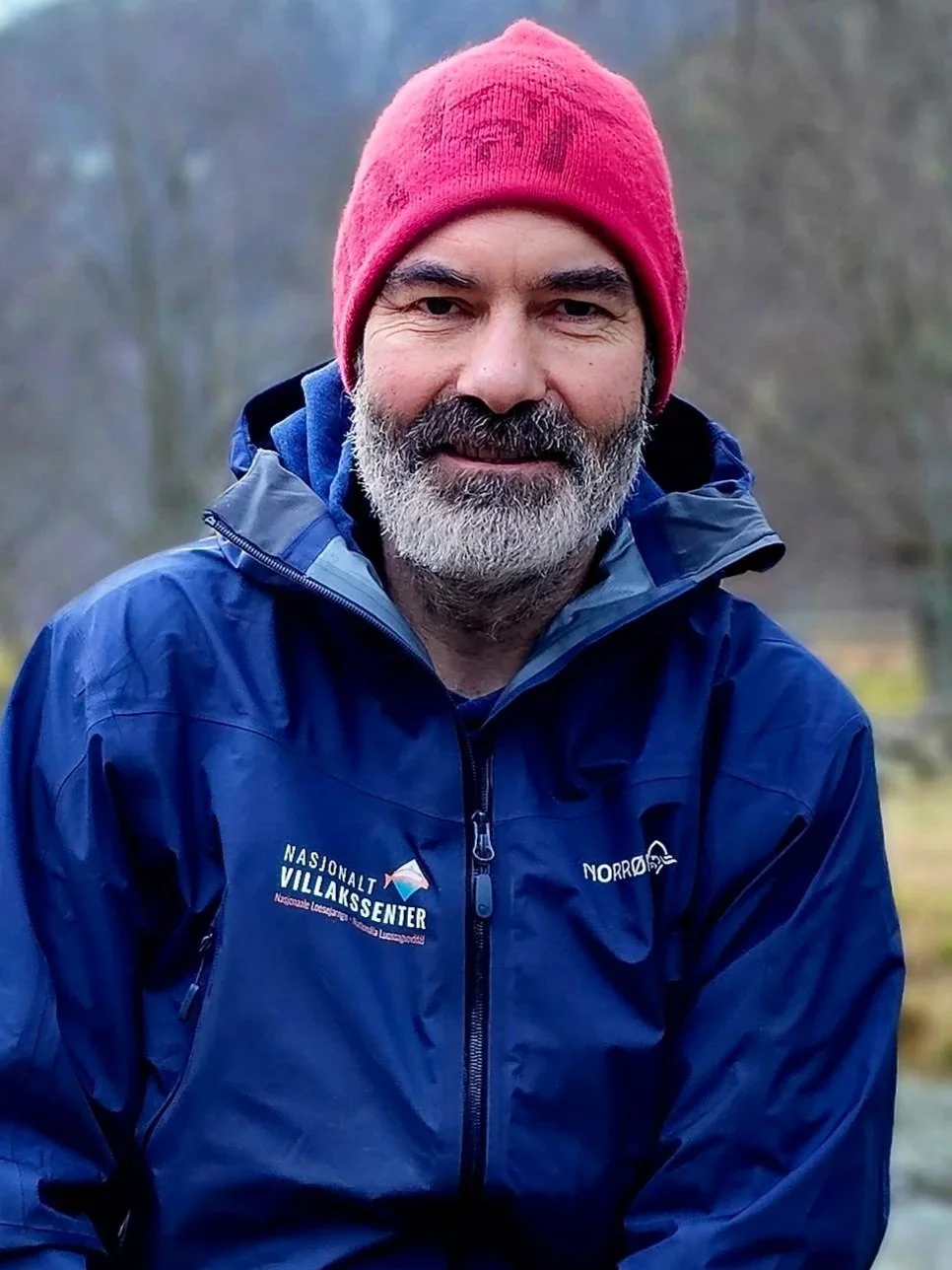 En mann med grått skjegg og måne bruker en rosa lue og blå jakke med logoer for Nasjonalt Villakssenter og Norre. Han står utendørs med trær i bakgrunnen.