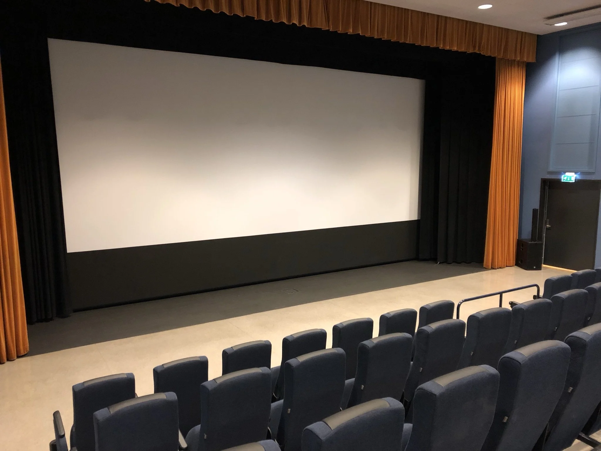 Foto av et kinolerret med svarte sideskjermer og oransje gardiner, med flere tomme stolrader foran.