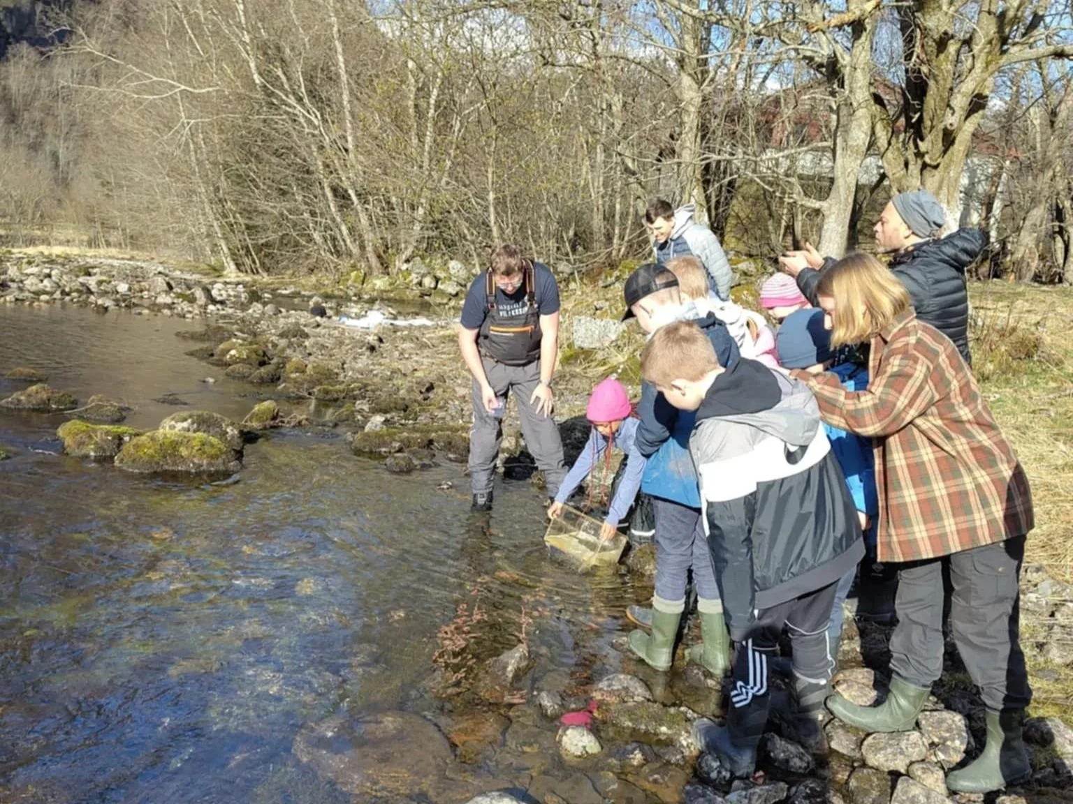 Unge utforsker langs en elv om våren, noen har på regnbukser og plukker noe med en bøtte.