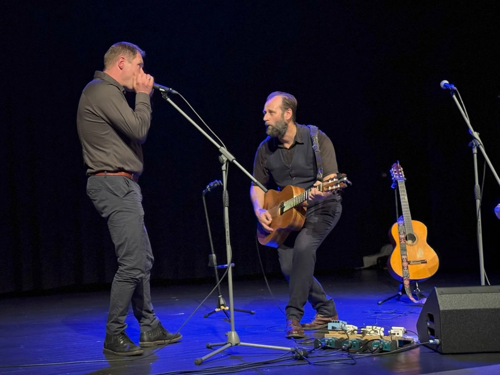 To menn spiller på scenen med to gitarer, en kaller og en mikrofon, med mørk bakgrunn.