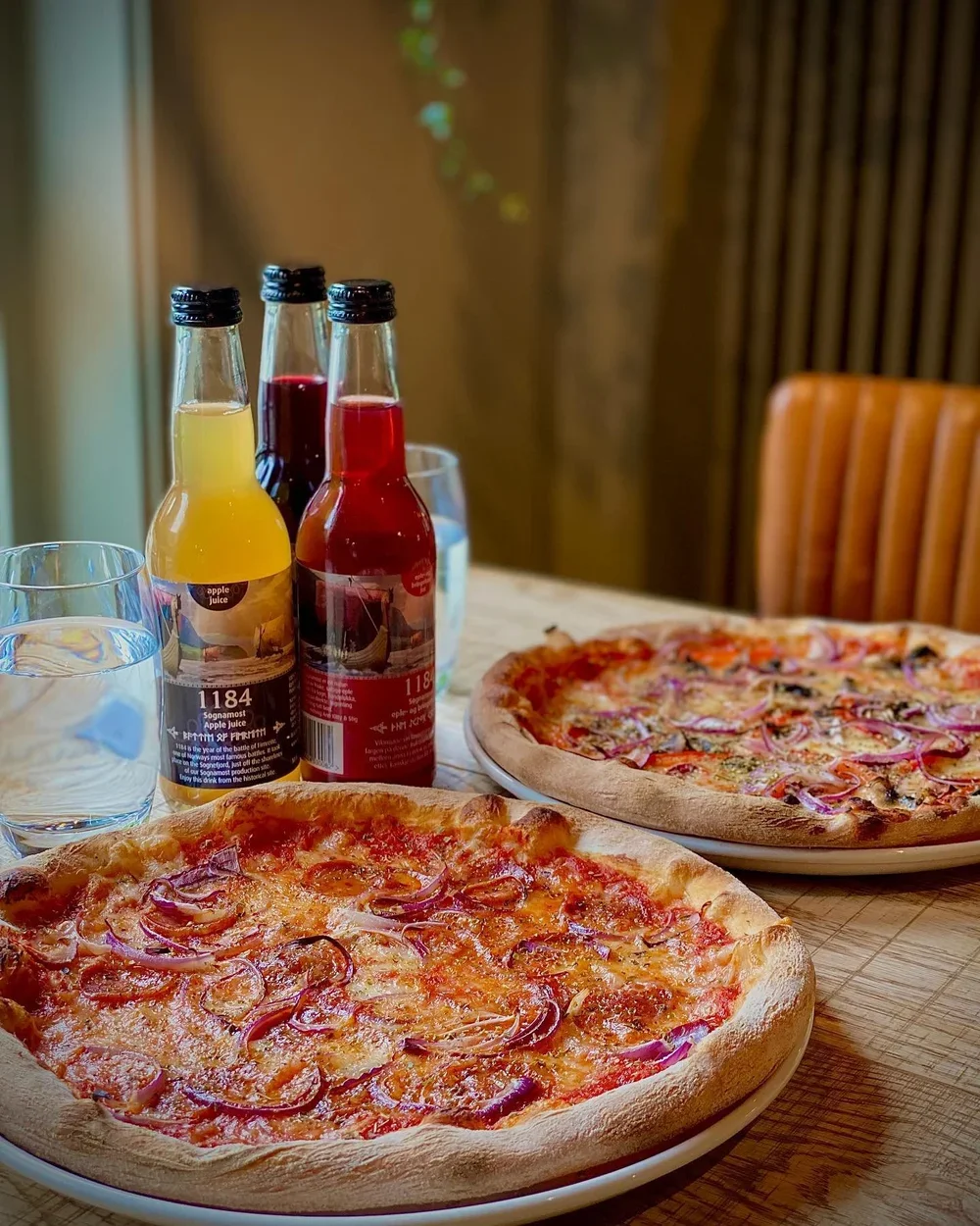 To pizzaer, ett med skinke og løk, på et trebord. Tre brusflasker og to glass med vann.