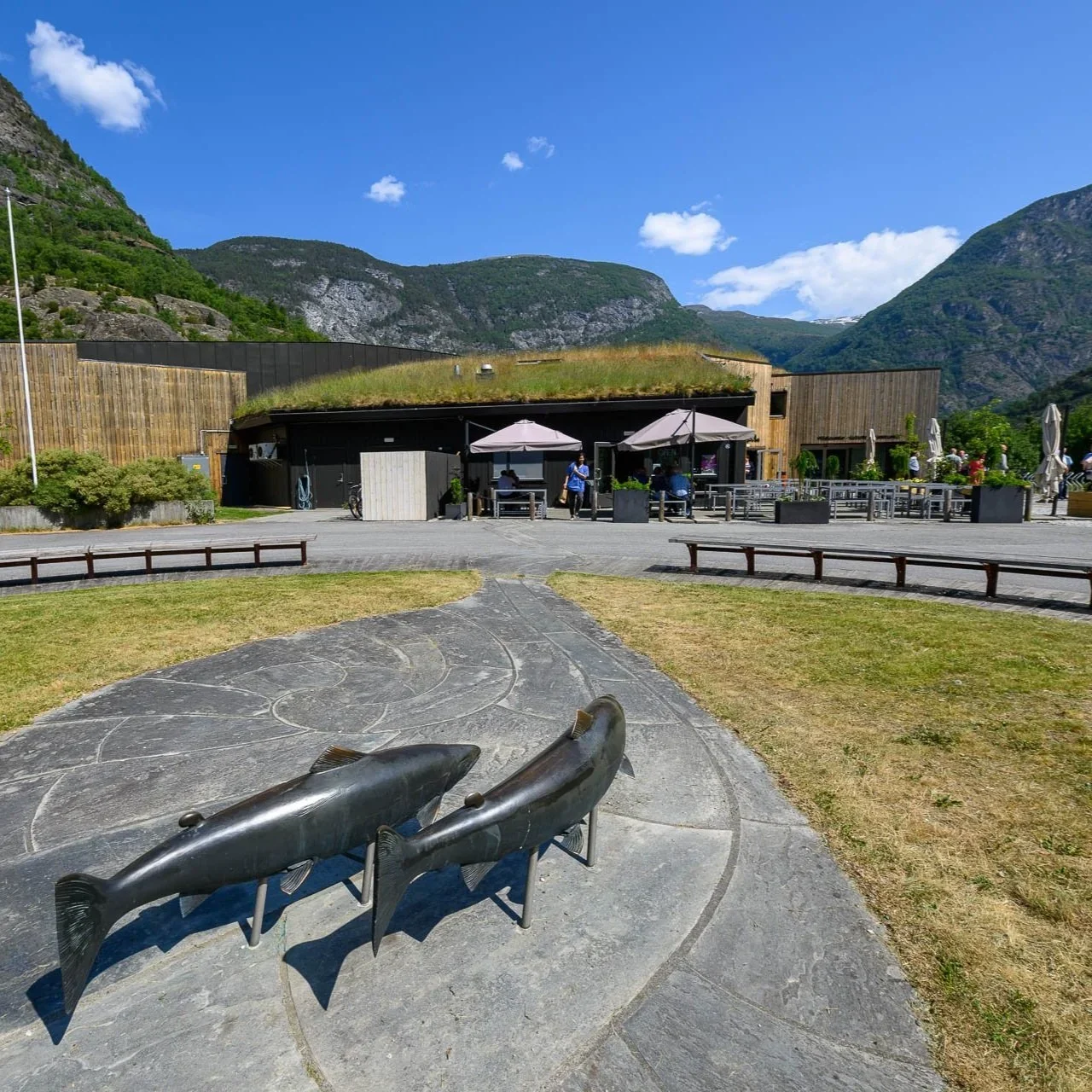 To store eller kafeer med utendørsterrasser, omgitt av fjell og grønt landskap, med to store fiskeskulpturer i forgrunnen.