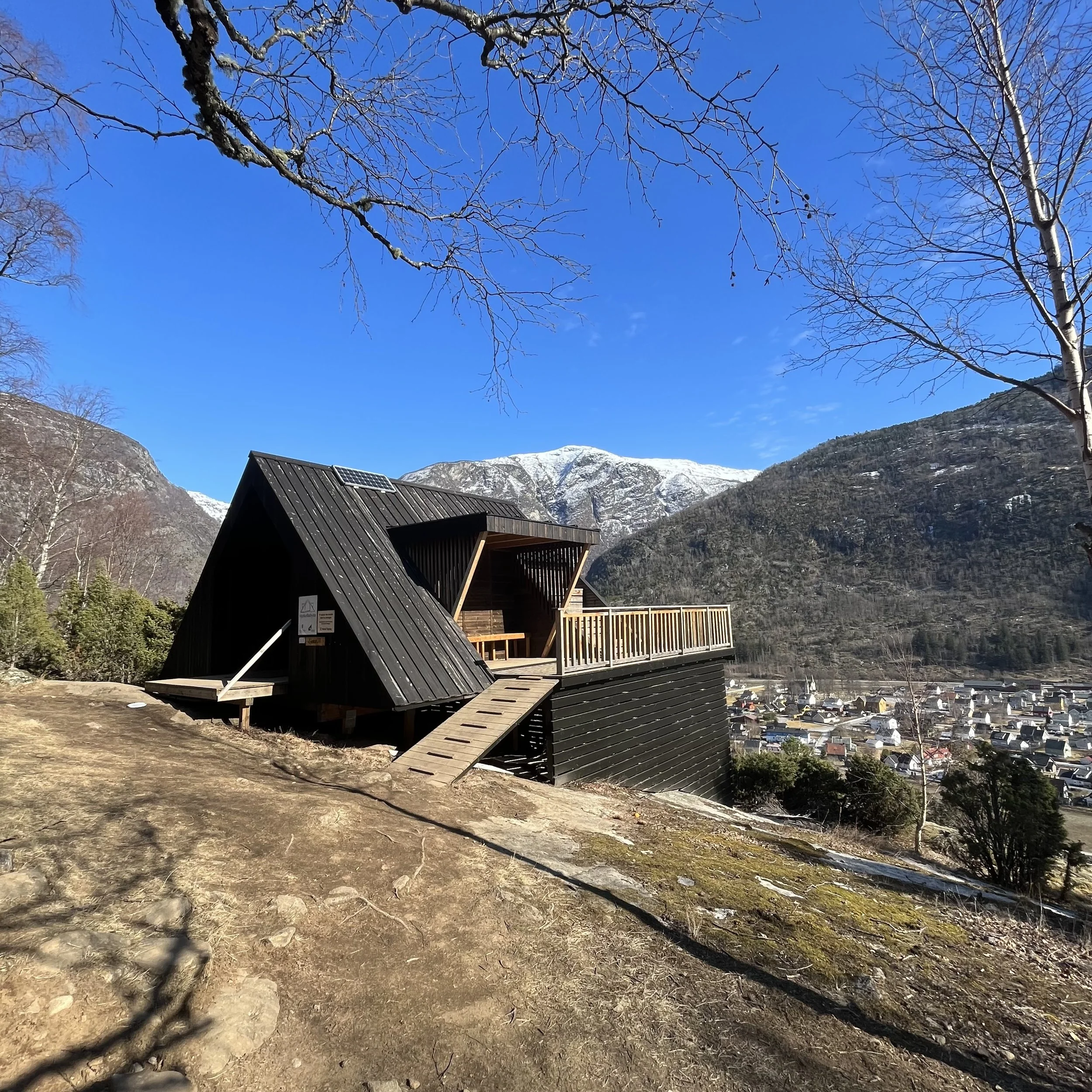 En moderne, svart tretakshytte med terrasse i fjelllandskap, omgitt av snødekte fjell og nesten nakne trær under en klar blå himmel.