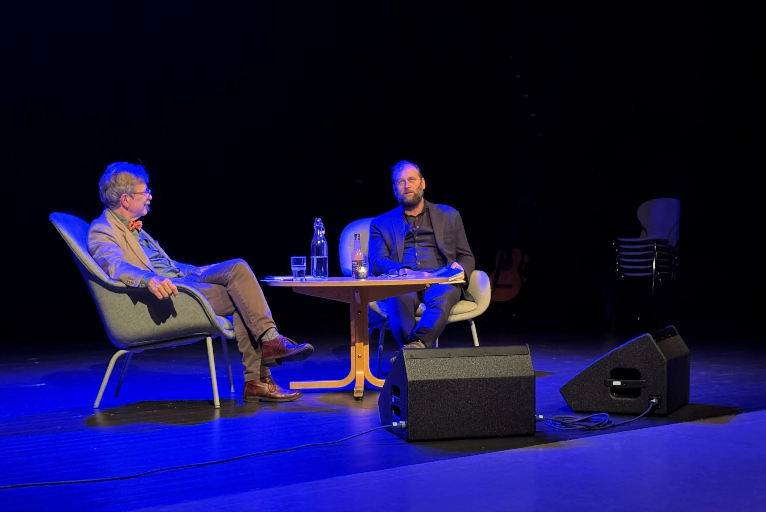 To menn sitter på en scene under en samtale. Den ene mannen har grått hår og briller, den andre har skjegg og mørk dress. Det er en gitar og to vannflasker på bordet mellom dem.