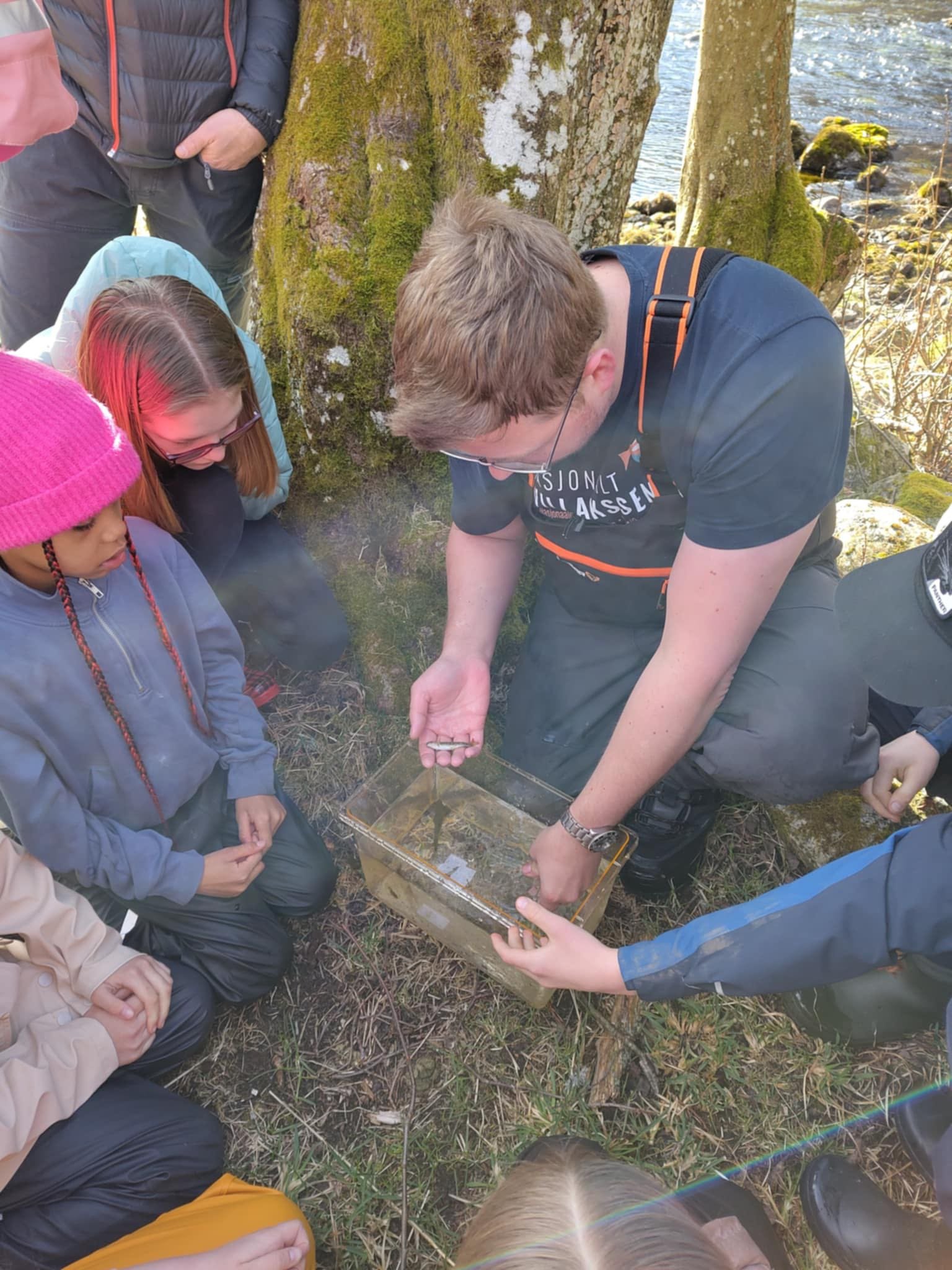 En gruppe mennesker, inkludert unge jenter og voksne, tett samlet rundt en vannfylling av en vannfelle eller ukjent utstyr, utenfor i et skogområde med trær og vann i bakgrunnen.