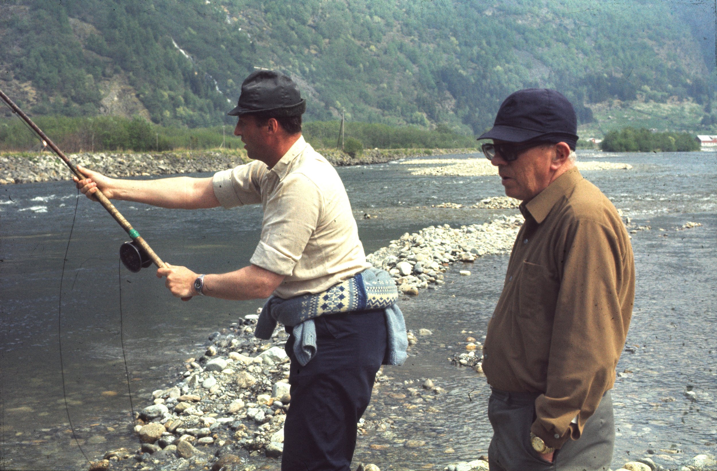To menn står ved elvekanten, den ene holder en fiskestang og er i ferd med å fiske, mens den andre ser på.