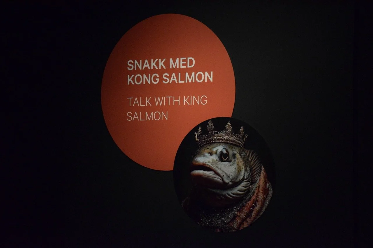 Informasjonstavle med tekst 'Snakk med kong salmon' og 'Talk with king salmon', samt et bilde av en laksehodet med en krone på hodet.