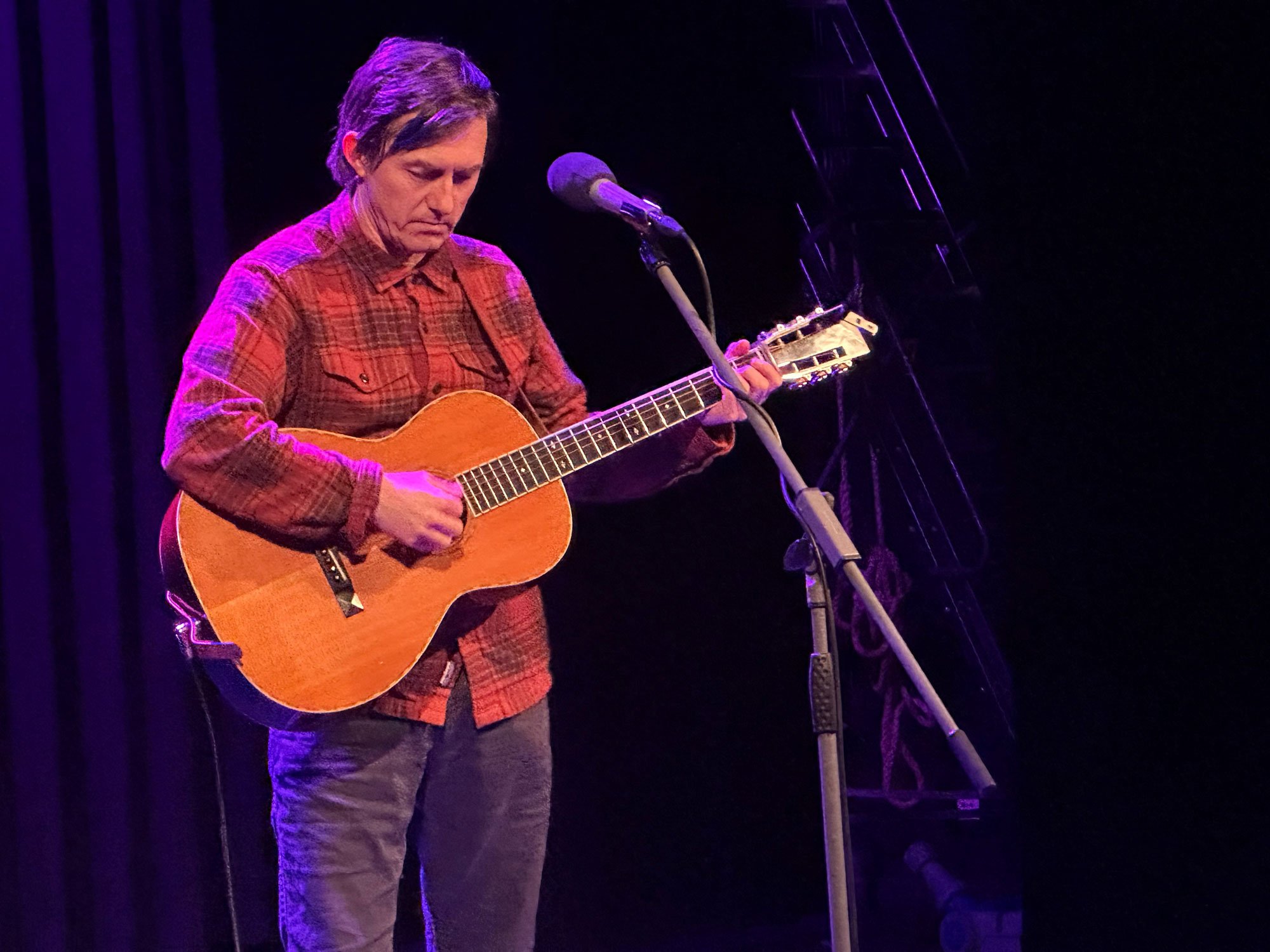 En mann spiller akustisk gitar på en scene med mørk bakgrunn og lys i lilla.