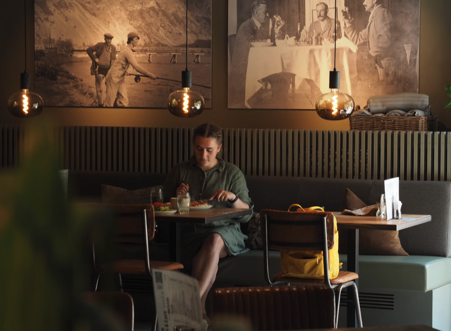 En kvinne som spiser alene på en kafé, med to store fotografier av folk i fortiden på veggen bak henne, og tre lysholdere som henger fra taket.