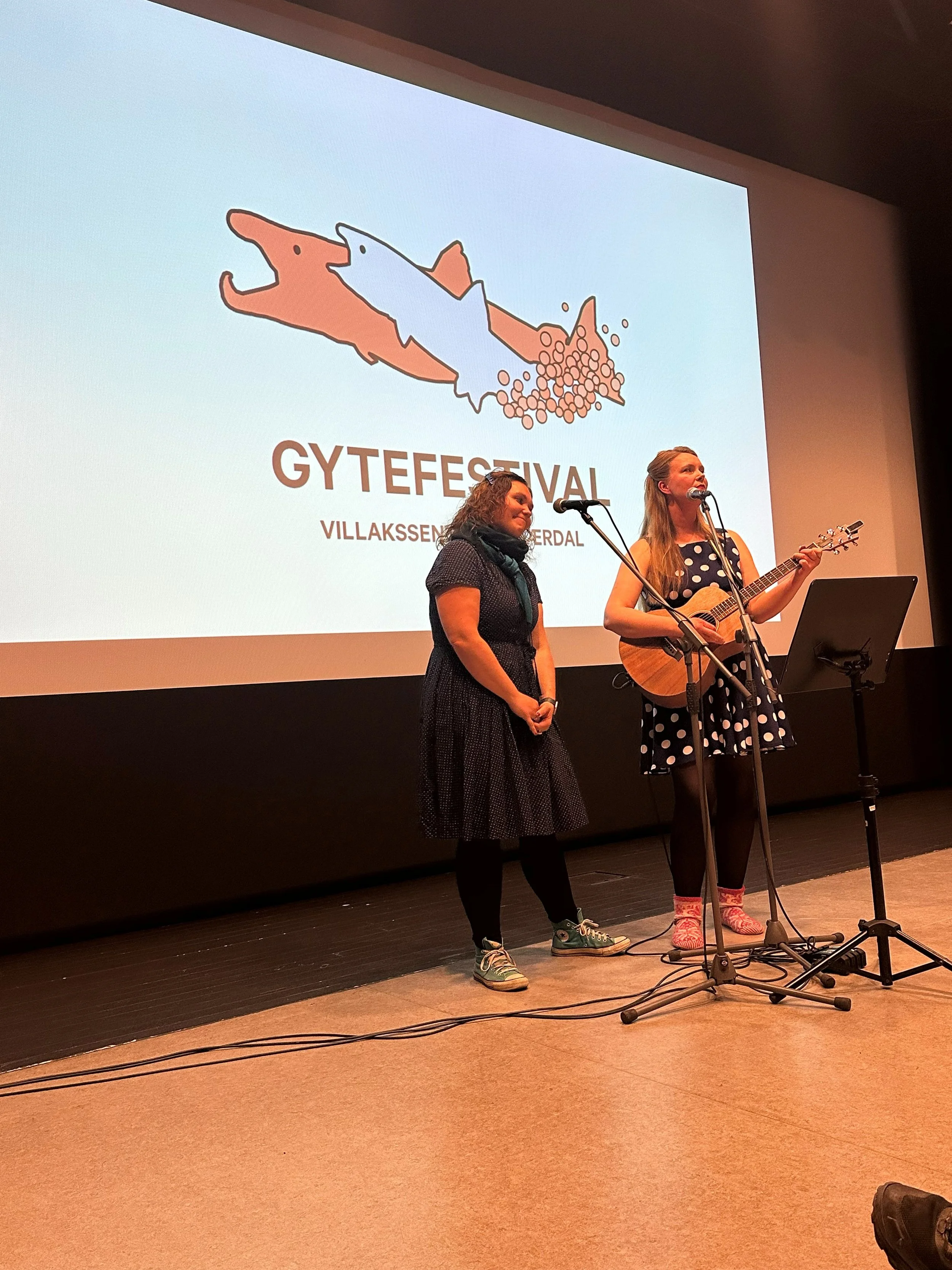 To kvinner står på scenen med mikrofoner. Den ene spiller gitar. Bak dem vises en stor skjerm med en logo for gytefestivalen, som viser to fisker som svømmer, en i en oransje farge og en i en blå farge, med teksten "GYTEFESTIVAL VILLAKSSEN-VALDERAL".