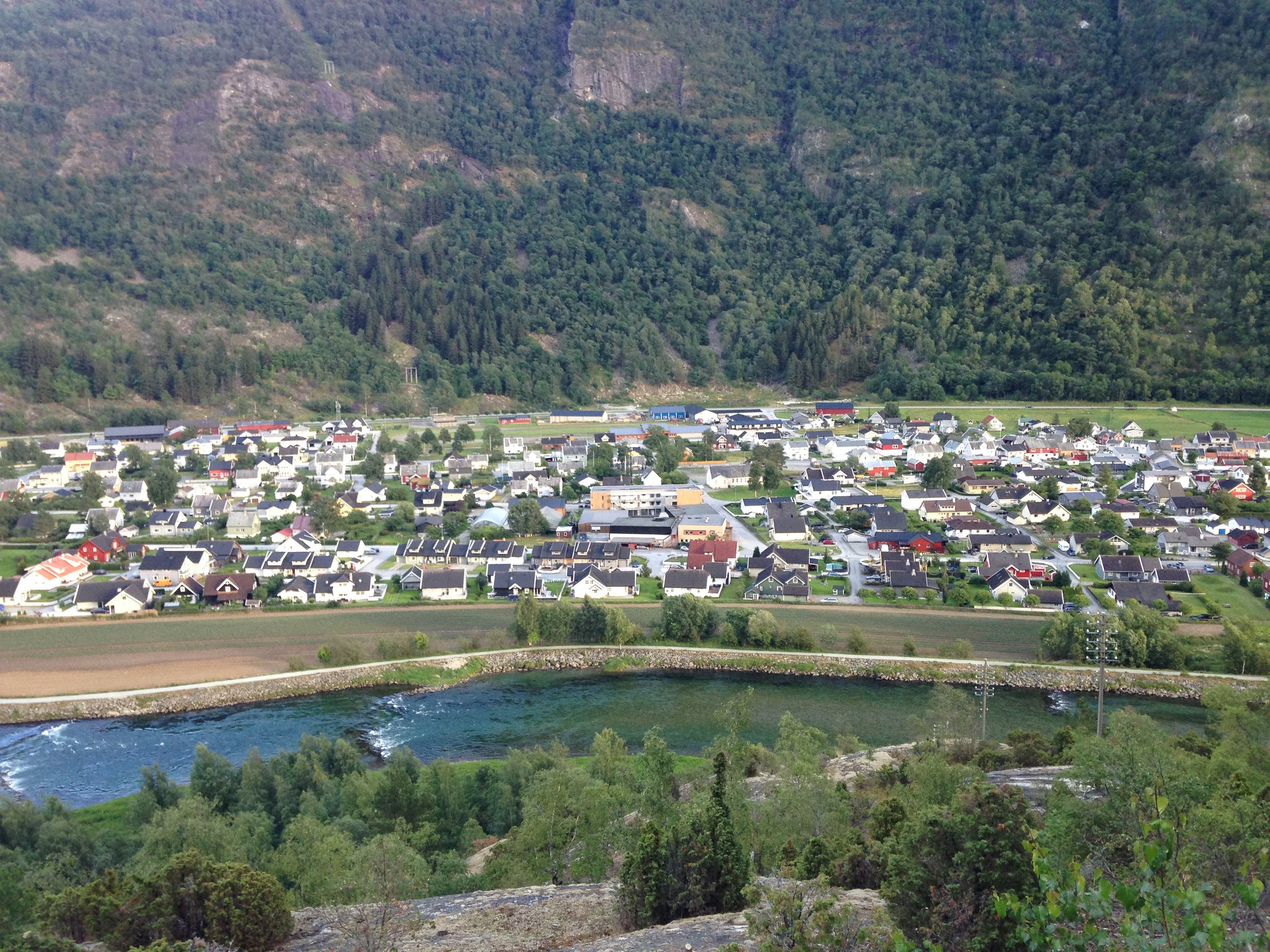 En liten by med en rekke hus ved en elv, omgitt av grønne åser og fjell.