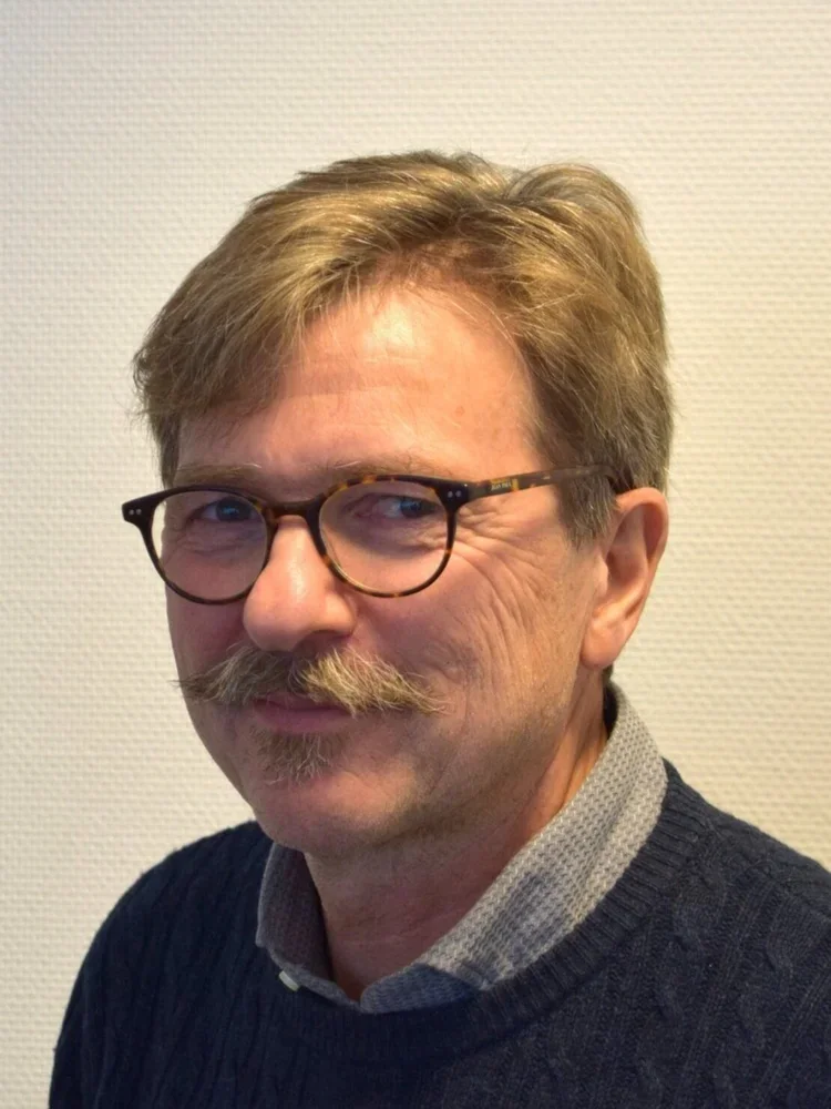 Et portrett av en mann med brunt hår, briller, bart og skjegg, iført en mørk genser over en grå skjorte, tatt innendørs mot en lys vegg.