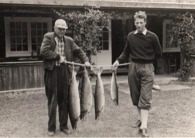 To menn holder en fiskestang med fire fisker, på en hage med hus i bakgrunnen.