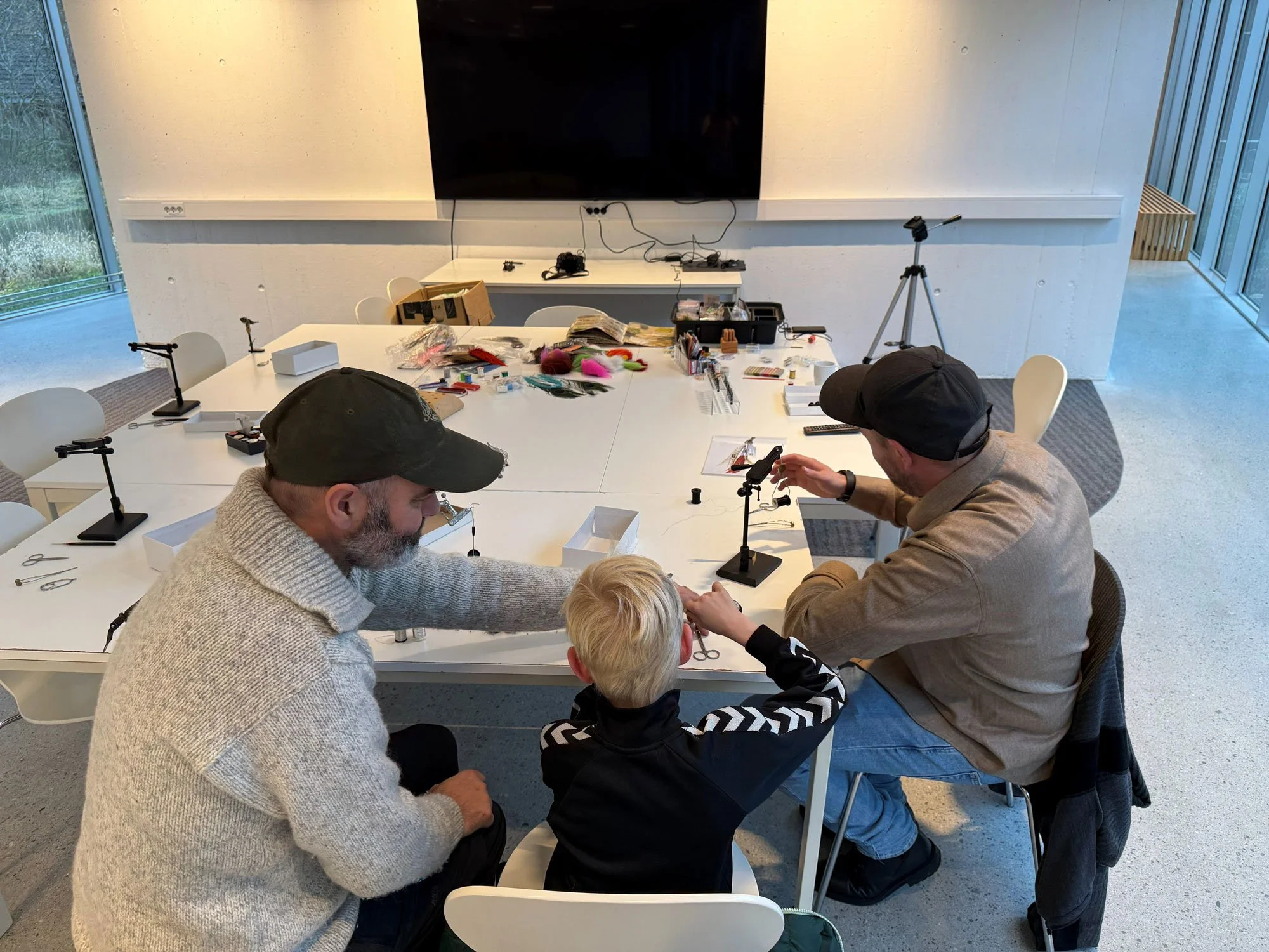 To voksne personer og ett barn som arbeider med fiskeutstyr i et rom med et stort bord og videovise på veggen.