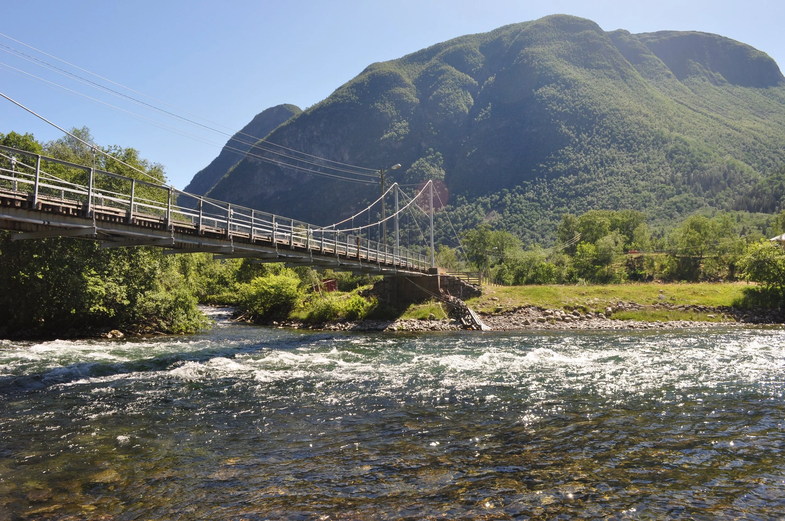 En bro over en elv med grønn skog og fjell i bakgrunnen på en solrik dag.