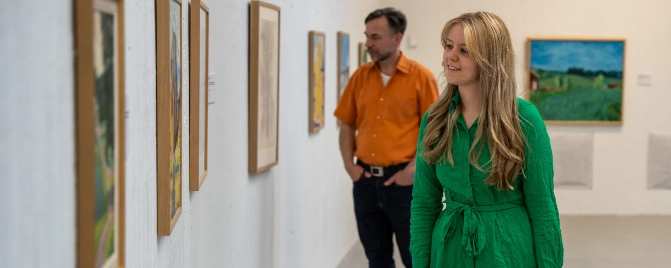 To personer ser på kunstutstilling i et galleri, en kvinne i grønn kjole og en mann i oransje skjorte.