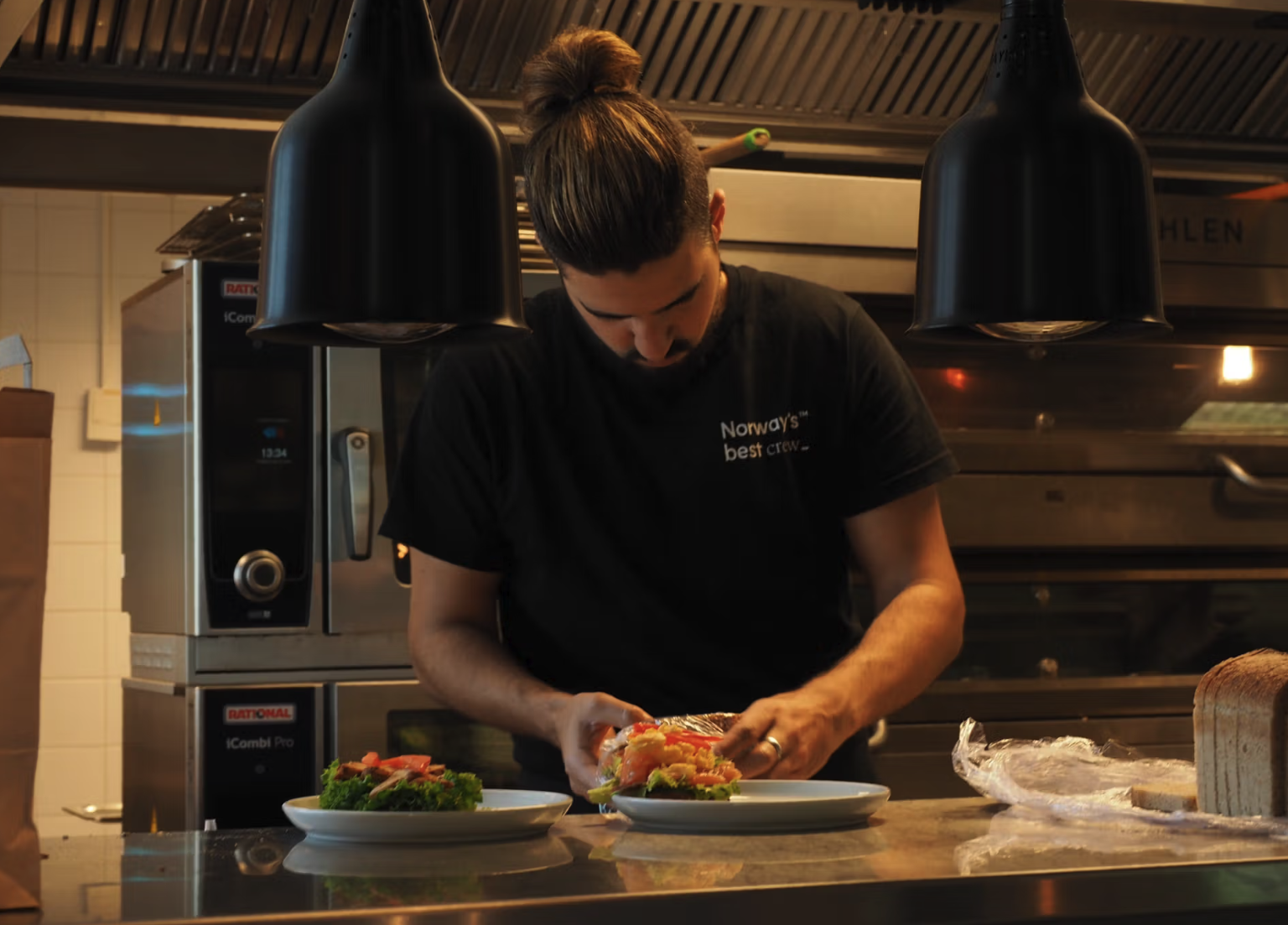 En mann med langt hår i hestehale lager mat i en profesjonell kjøkken. Han har på seg en svart T-skjorte som sier 'Norway's best crew'.