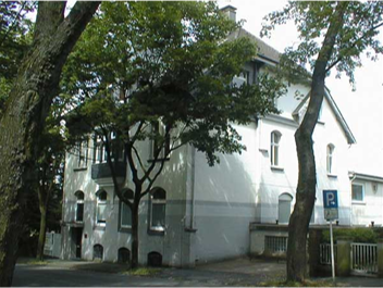 Ein weißes Mehrfamilienhaus mit mehreren Ebenen, umgeben von Bäumen in einer Wohnstraße.