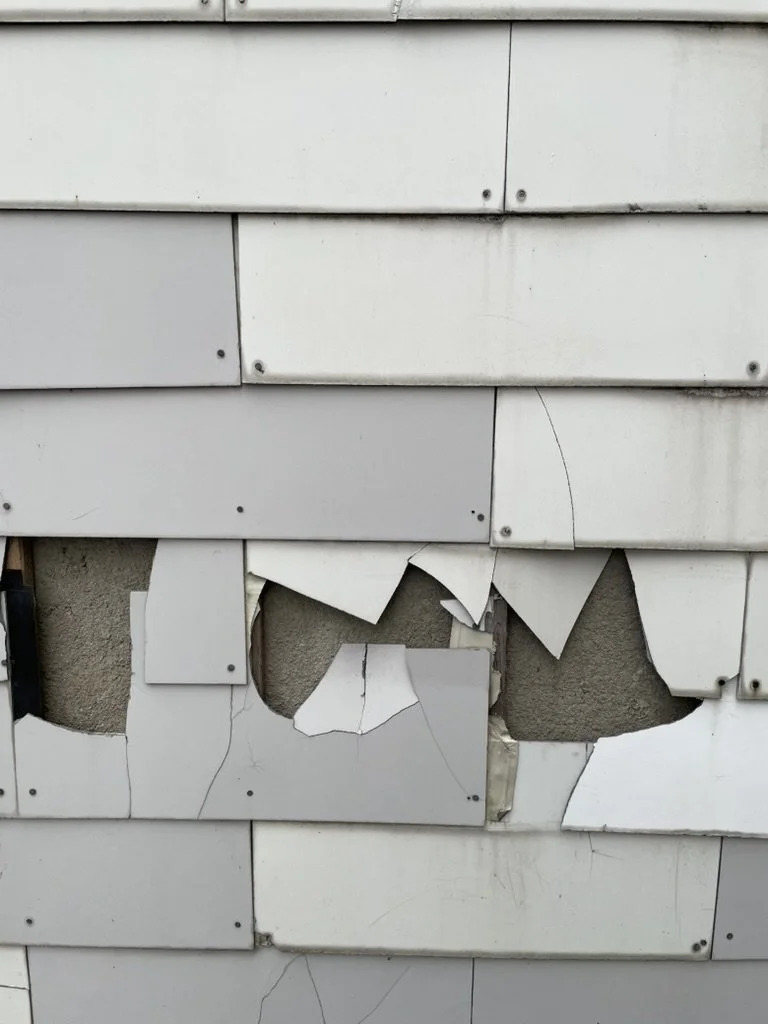 Damaged gray siding on a building with several cracks, missing and loose pieces, exposing the underlying wall.