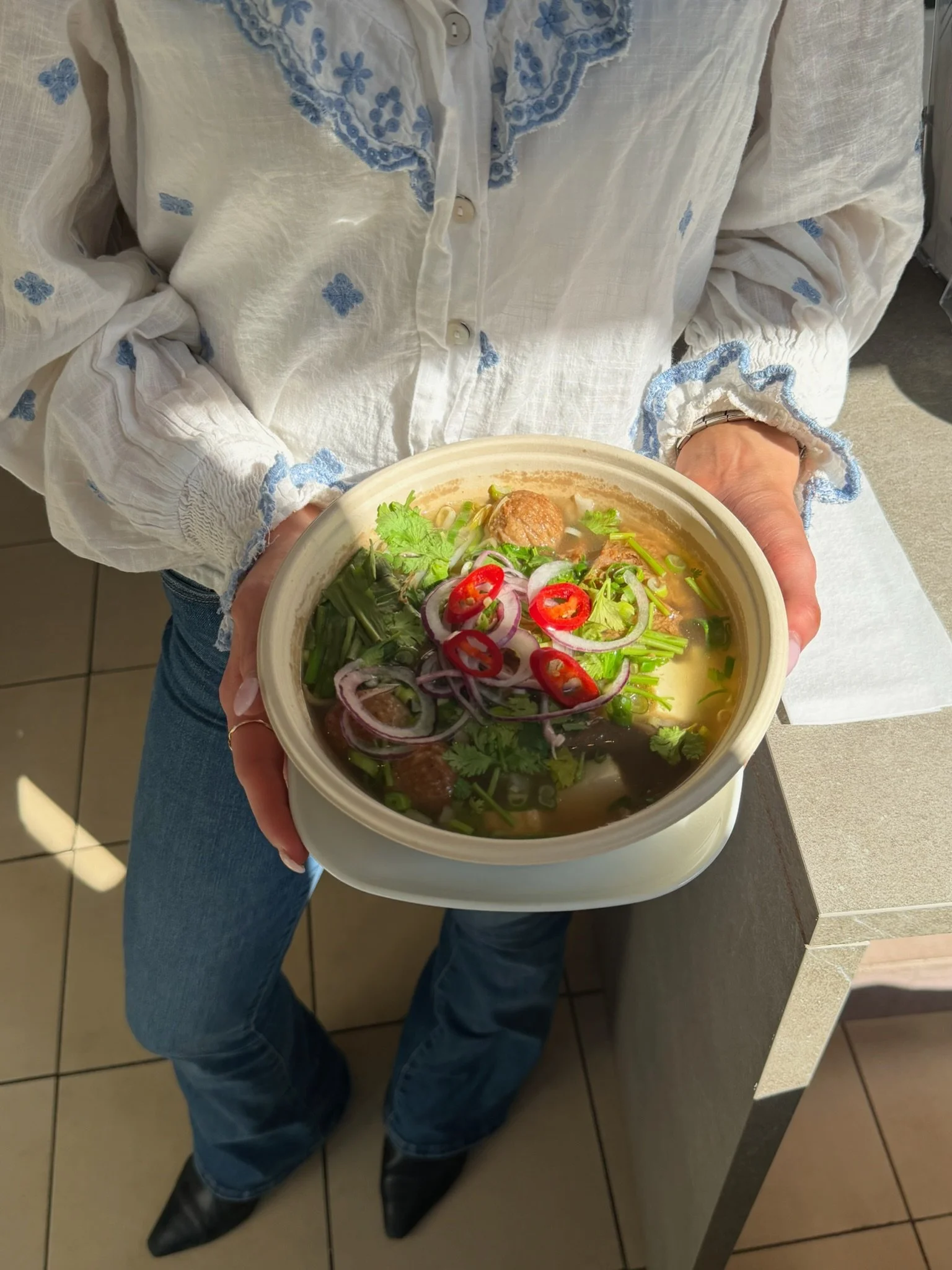 En person i blå jeans og svart støvel holder en bolle med suppe, fylt med grønnsaker og veganske kjøttboller, servert på et serveringsbord.