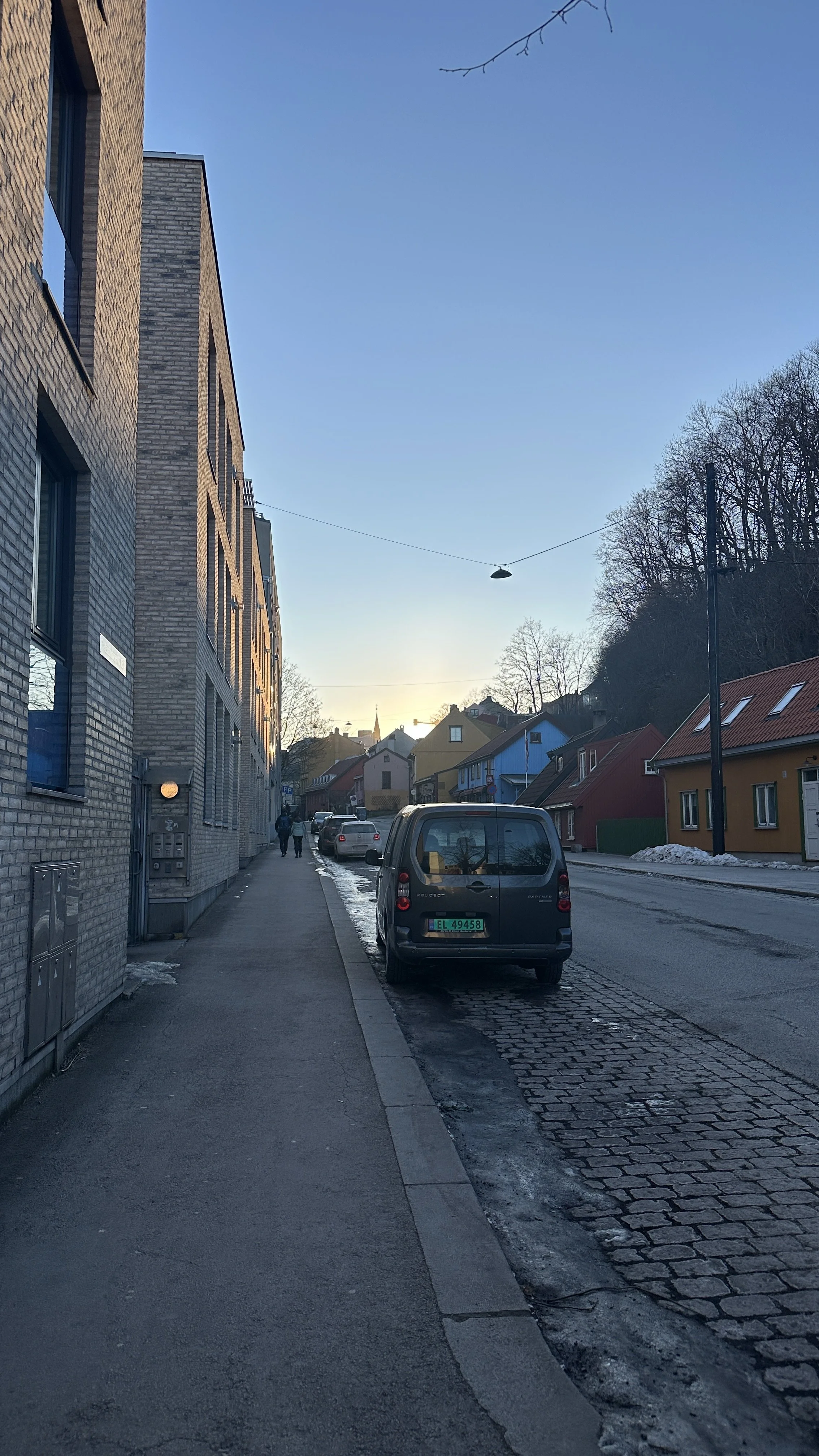 En gate i en by med parkerte biler, fargerike hus og en klokkespill i bakgrunnen. Det er vinter med snø på fortauet og i veikanten.
