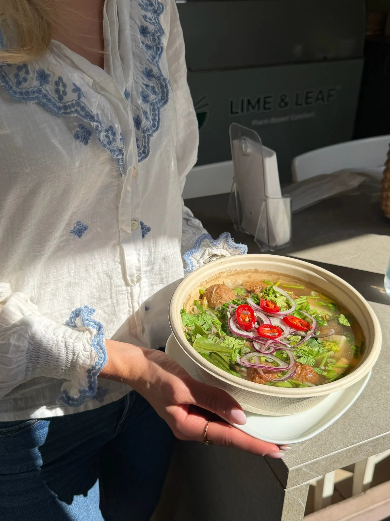 Person holding a bowl of Pho soup with vegetables, vbegan meatballs and chili.