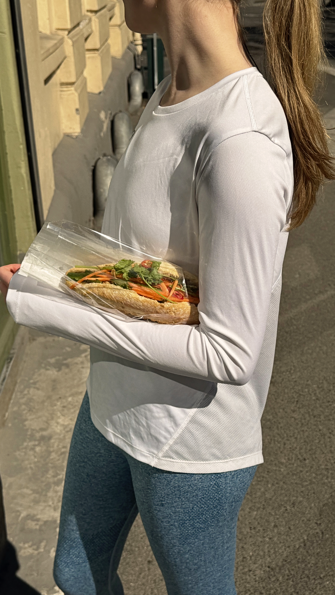 A woman in white long-sleeved workout clothes holds a plastic-wrapped baguette with salad and vegetables.
