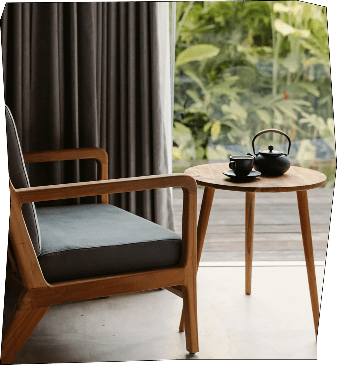 A cozy corner with a wooden armchair featuring a dark cushion, next to a round wooden table holding a black teapot and cup, with a window revealing green foliage outside.