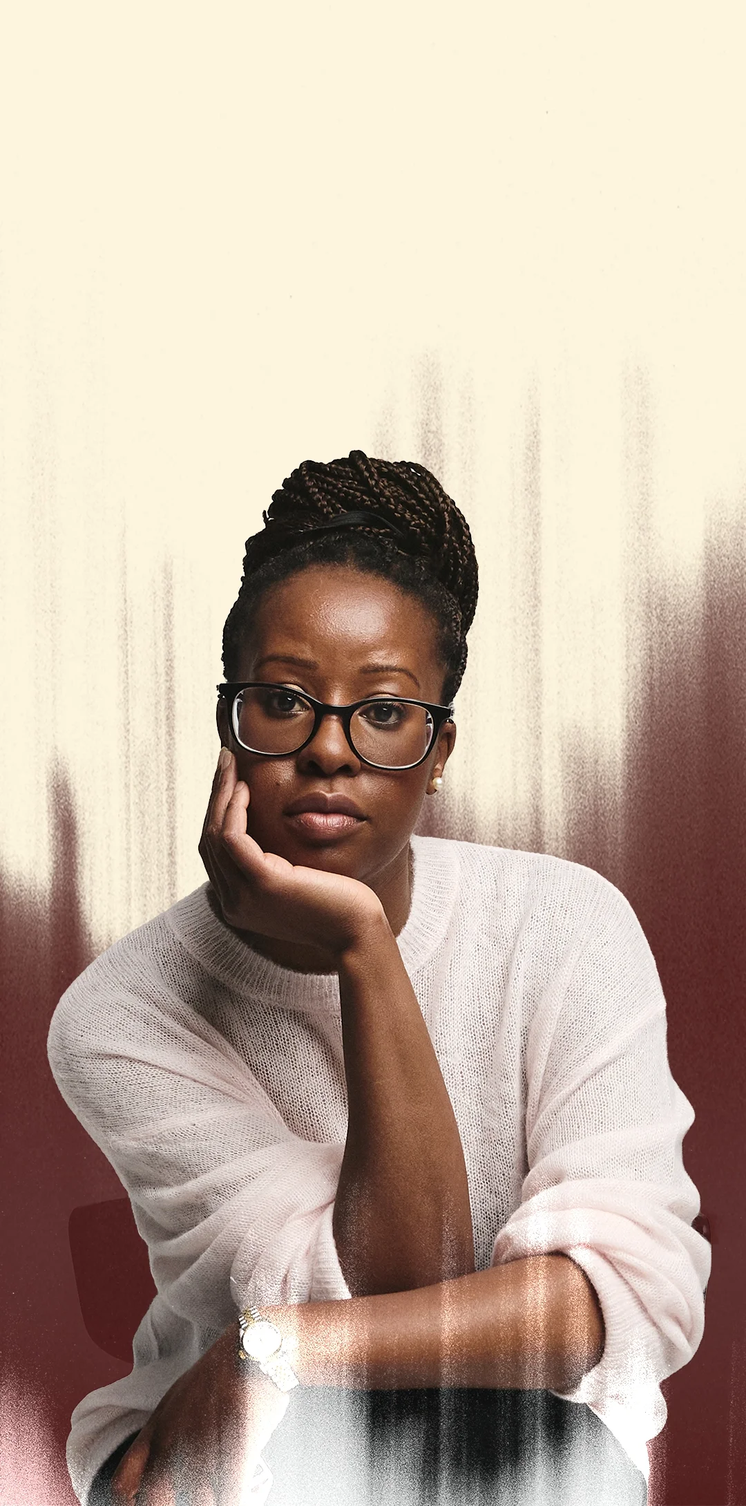 A woman with glasses and braided hair, resting her chin on her hand, wearing a white sweater, sitting in front of a plain background.