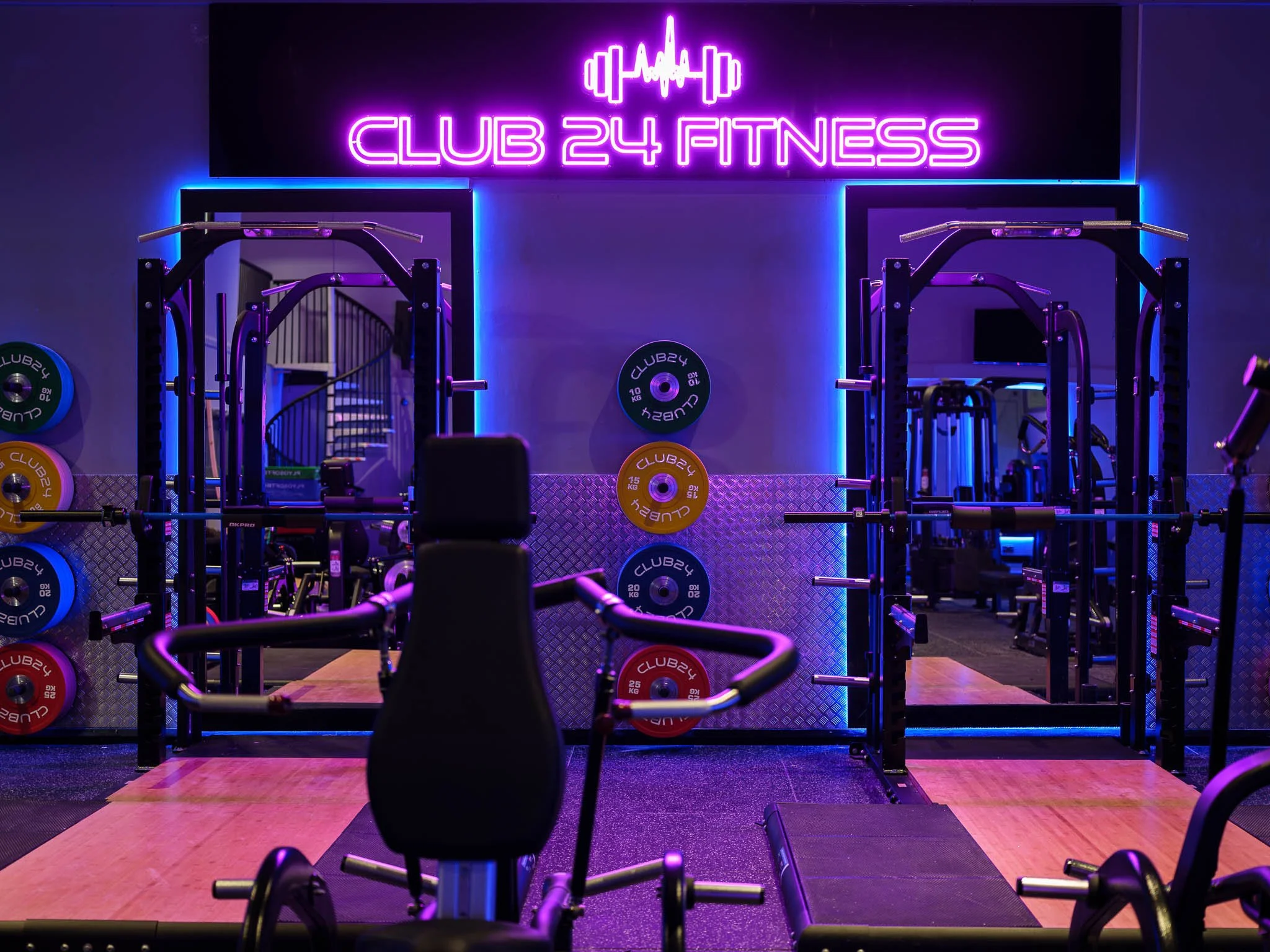 An empty fitness gym with weightlifting equipment and neon sign that reads 'Club 24 Fitness.'