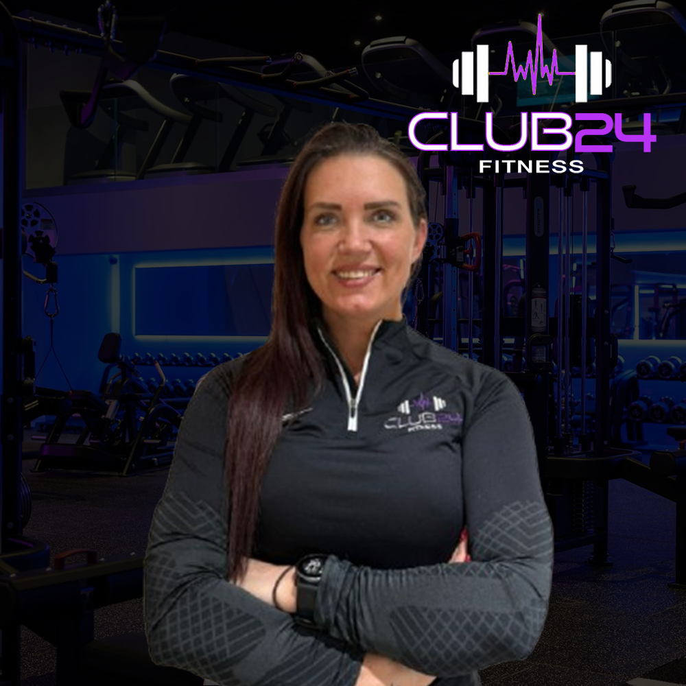 A woman smiling with arms crossed wearing a black athletic jacket with the CLUB24 FITNESS logo, standing inside a gym with exercise equipment and a sign that reads CLUB24 FITNESS.