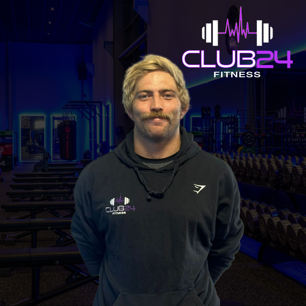 A man with blonde hair and a mustache standing inside a fitness gym. He's wearing a black hoodie with a "Club 24 Fitness" logo on the front, and is smiling slightly. The gym background shows weight racks and exercise equipment, with a large illuminated sign that reads "Club 24 Fitness" featuring a barbell and heart monitor lines.