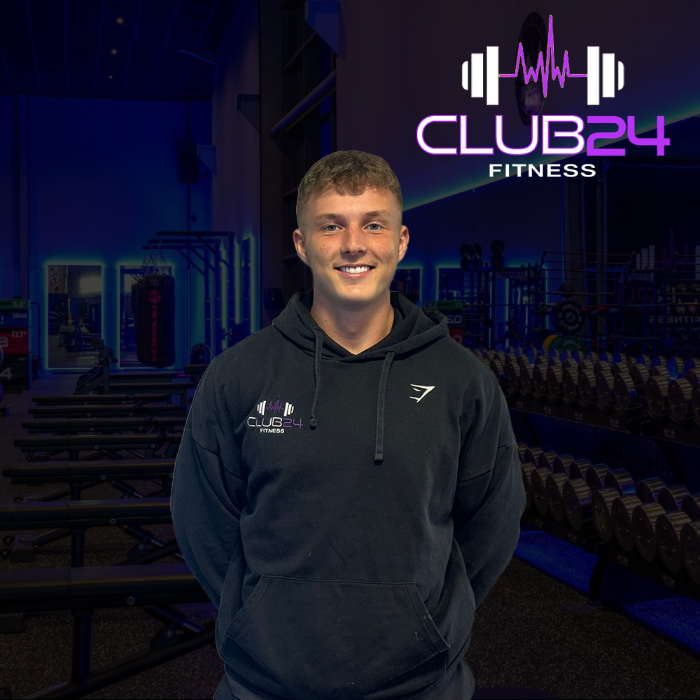 A young man smiling stands in a gym with weights and fitness equipment. Behind him is a sign that reads 'Club 24 Fitness' with a logo of a barbell and sound wave. He is wearing a black hoodie with the same logo.