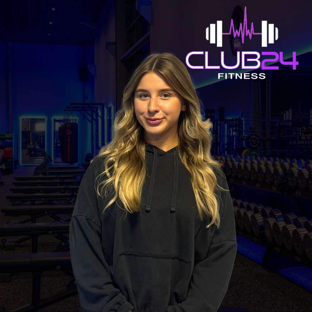A young woman with long blonde hair standing in a fitness gym. The gym has workout equipment and a neon sign that reads 'CLUB24 FITNESS' with a barbell and sound wave graphic.