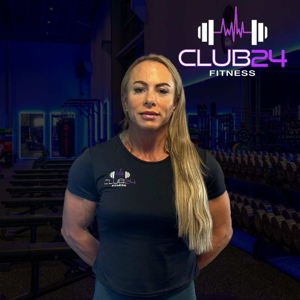 A woman standing in a gym with a 'Club 24 Fitness' logo on her black t-shirt, in front of a wall with the same logo and various gym equipment including dumbbells and punching bags.