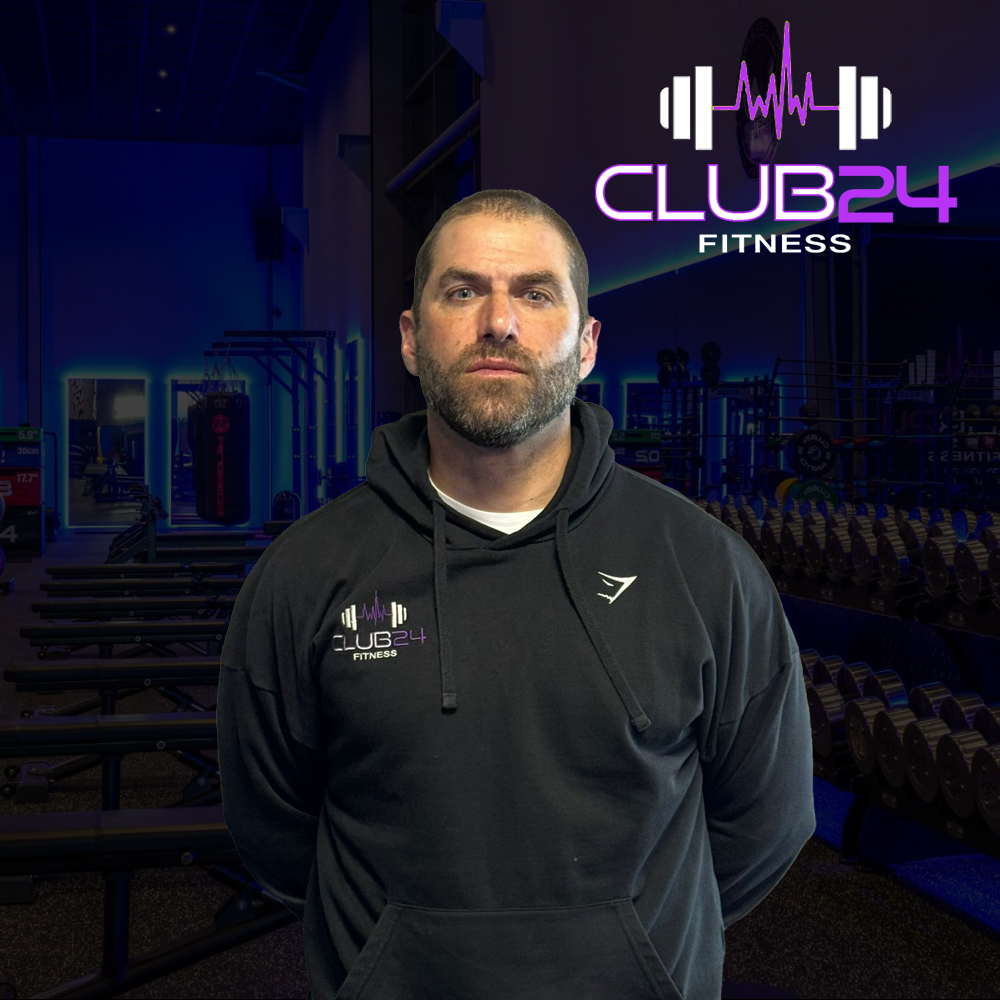 A man wearing a black hoodie with 'Club 24 Fitness' logo stands inside a gym with weightlifting equipment and a neon sign with the same logo in the background.