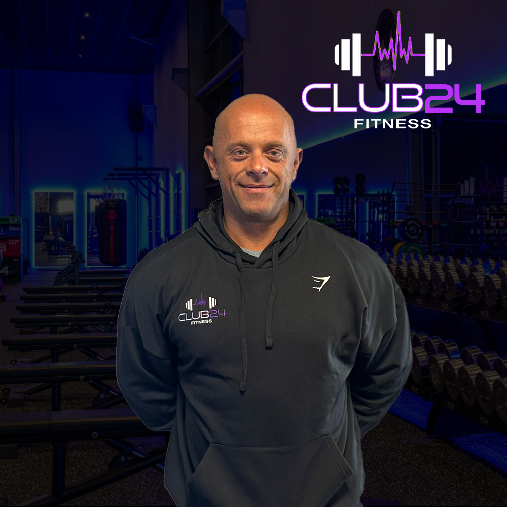 A man in a black hoodie stands in a fitness gym with weightlifting equipment in the background. The hoodie and a wall display show the logo 'Club 24 Fitness' with a purple and white color scheme, and the background features a dark, modern gym interior with neon lighting.