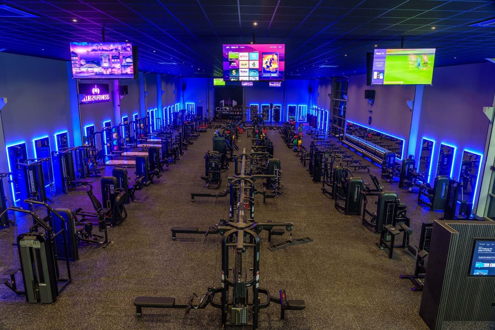 Empty gym with workout equipment, blue neon lighting, and large screens on the wall.