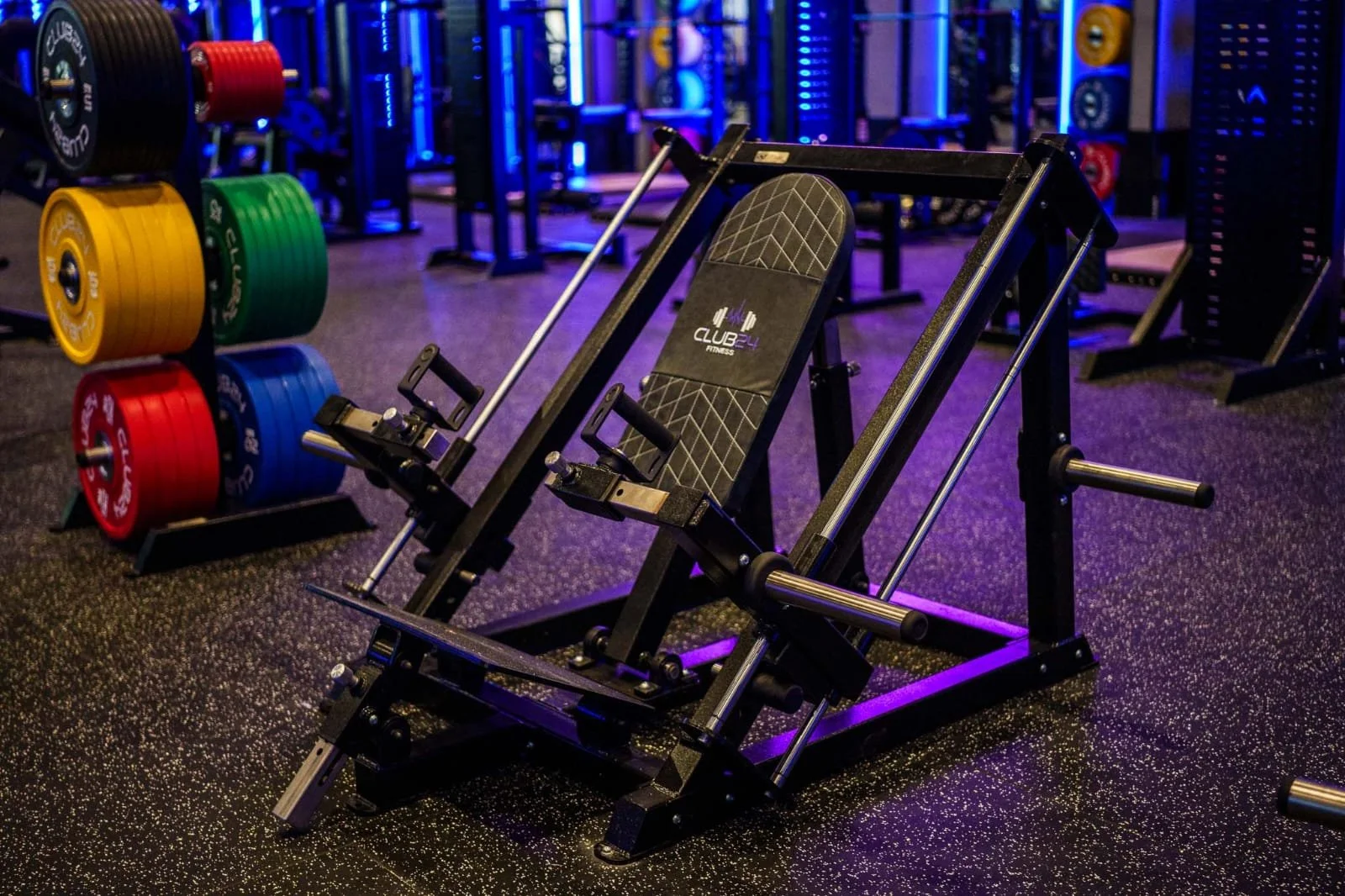Gym equipment in a fitness center, featuring an inclined sit-up bench and weight plates organized on a rack, with purple and blue accent lighting.