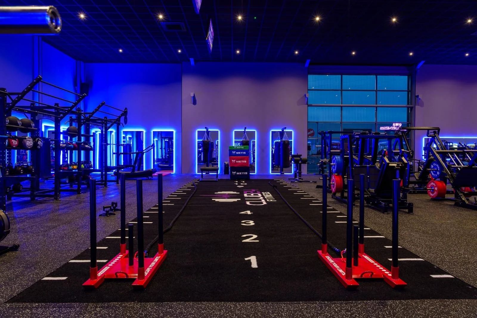 Empty fitness gym with black carpet featuring white numbered lanes, blue neon lighting along walls, and various workout equipment including weights, punching bags, and training stations, with a large window in the background.